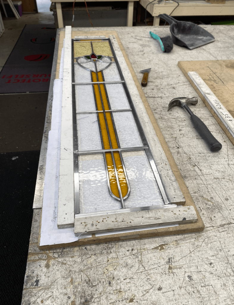 A stained glass panel featuring yellow and green designs, placed on a workbench with tools and materials around it.